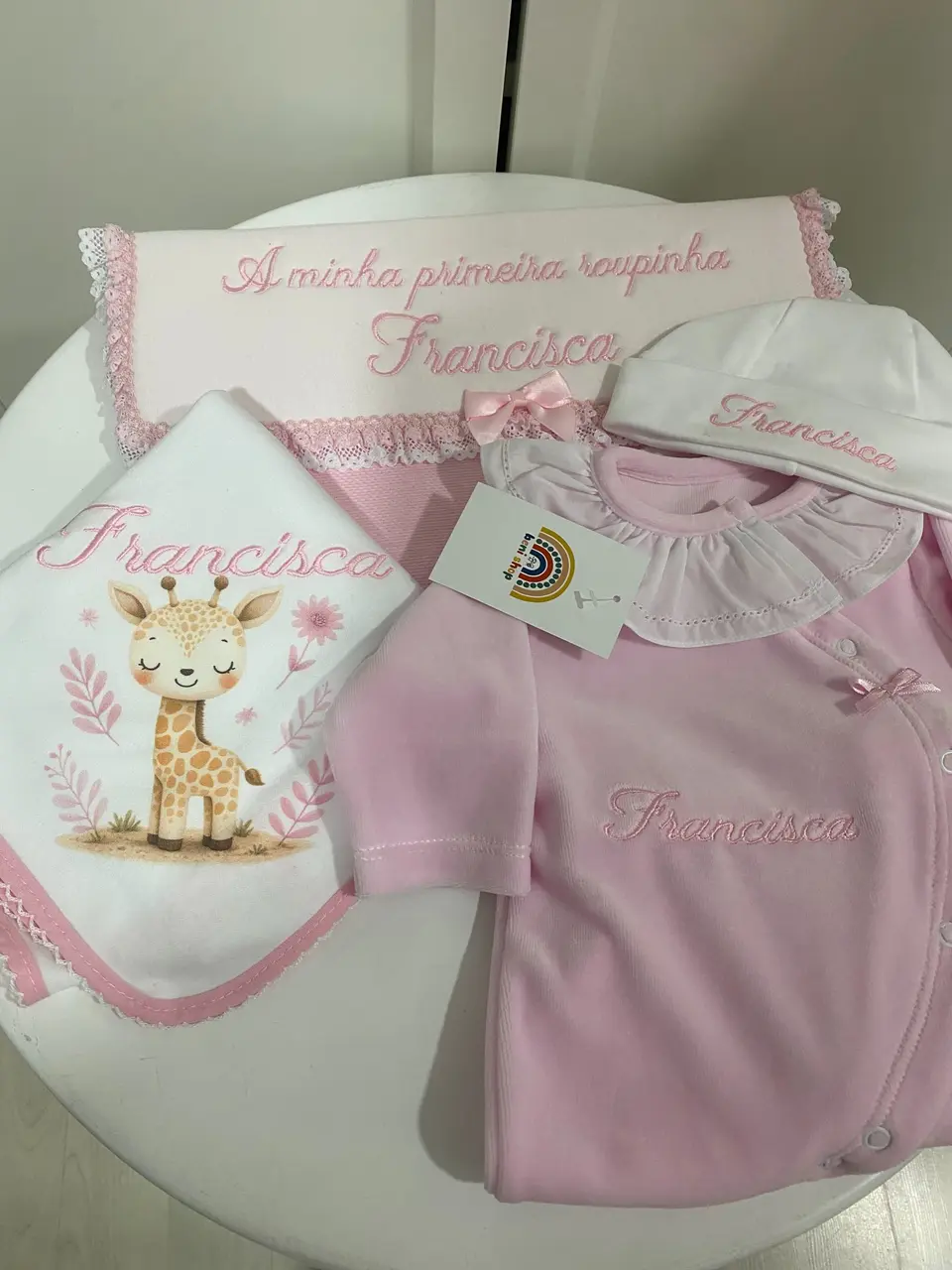A primeira roupinha da Francisca: babygrow rosa, gorro e toalha com o nome bordado e uma girafa.
